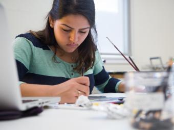 Student sits at workstation painting, surrounded by art supplies